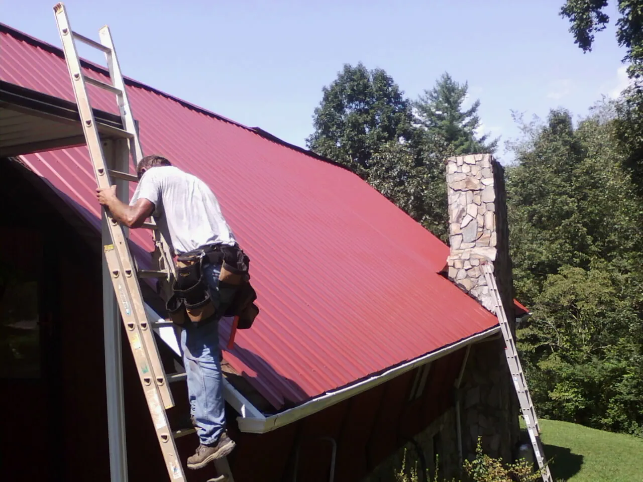 Roofer on a ladder beside a red metal roof and stone chimney — Soffit & Fascia Repair by Roofing Brothers in Spring Run